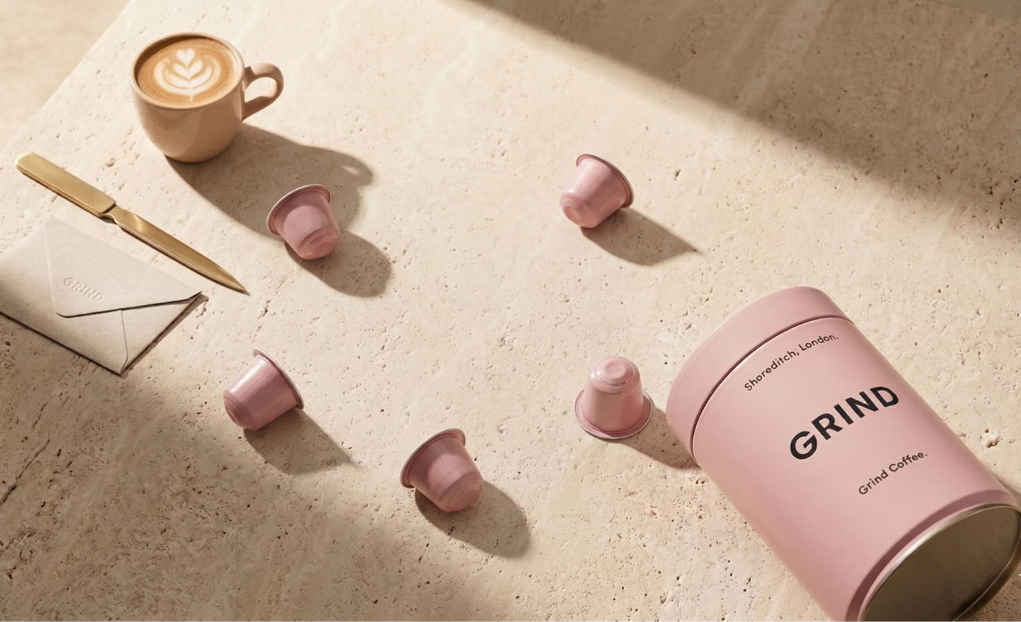 Minimalist coffee setup with a pink GRIND tin, scattered pods, and a latte under soft sunlight.