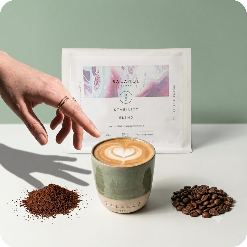 A hand points toward a white bag of Balance Coffee Blend behind a ceramic cup of latte art coffee, with a small pile of ground coffee and a small pile of whole coffee beans on a white surface against a pale green background.
